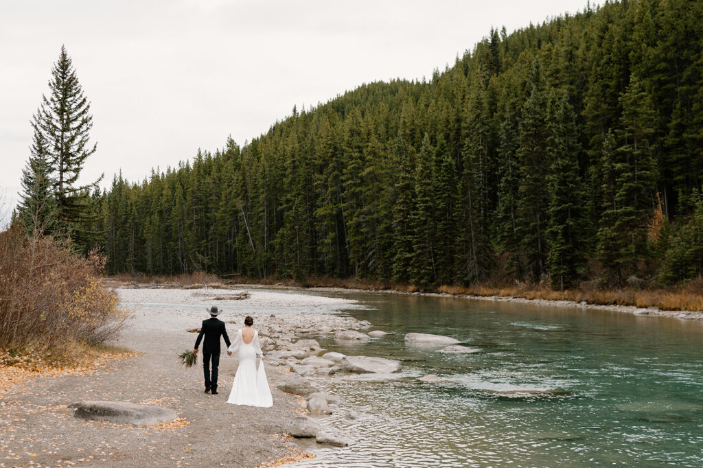 A couple walks hand and hand along the shoreline of an alpine stream near Lake Louise as they elope in Banff. 