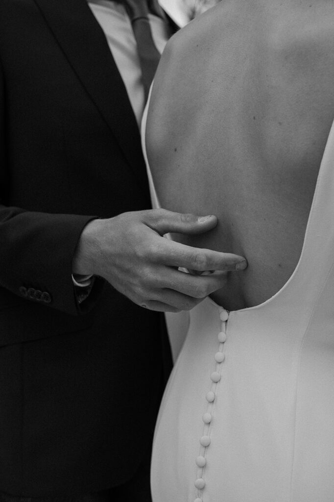 A man lightly touches his brides back, who is wearing a button back wedding gown. 