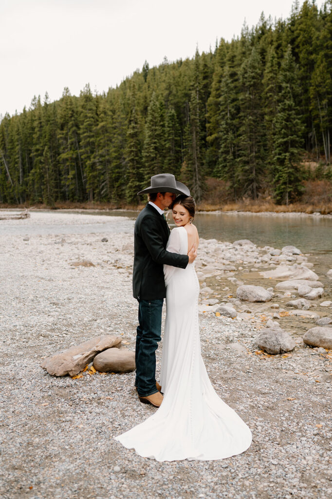 A man kisses his bride on the head near a stream at Lake Louise. 