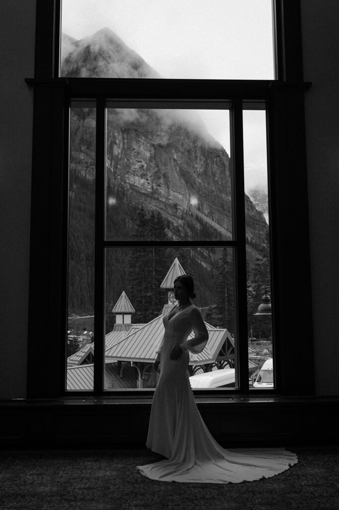 A bride in a long wedding gown poses in front of a large, floor to ceiling window at the Fairmont Chateau Lake Louise.