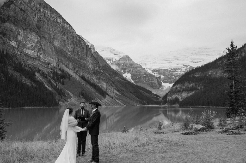 A bride and groom exchange vows lakeside in Banff while their officiant, Married By Cole stands in the background. 