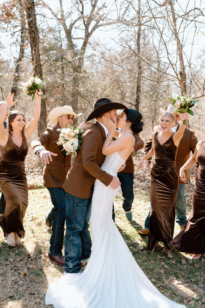 A bridal party cheers with groomsmen in cowboy hats as the bride and groom kiss in celebration of their rustic microwedding. 