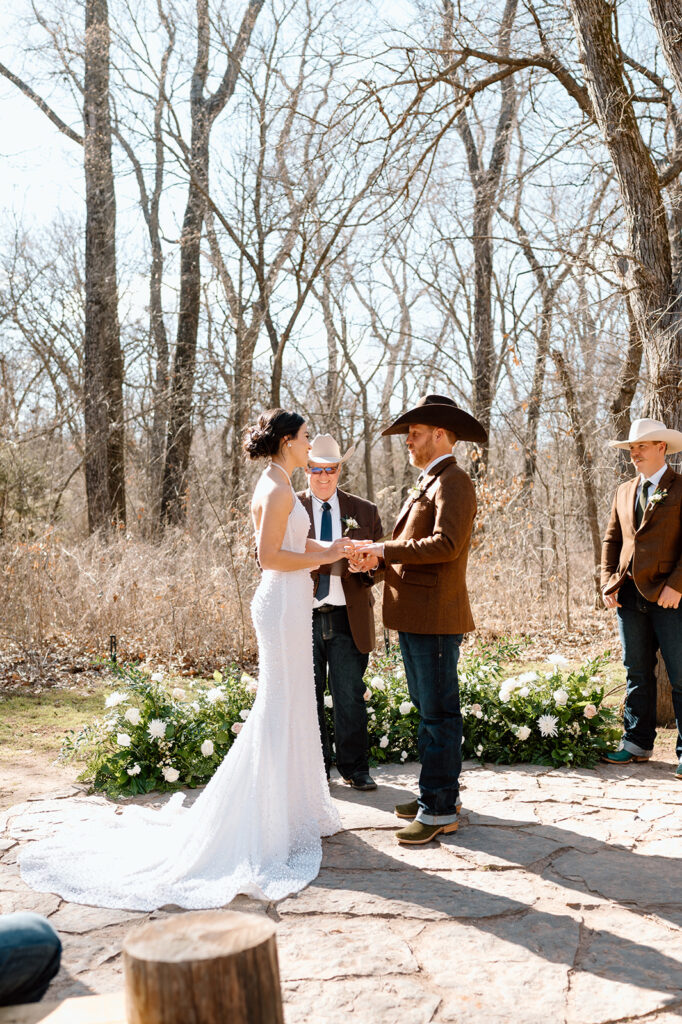 A man in a cowboy hat and brown blazer officiates a rustic microwedding for a bride and groom. 