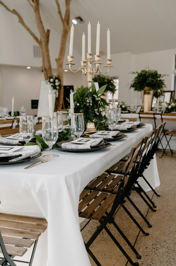 A microwedding tablescape with a white tablecloth, gold-rimmed glasses, gold cutlery and a printed-napkin place setting near taper candles. 