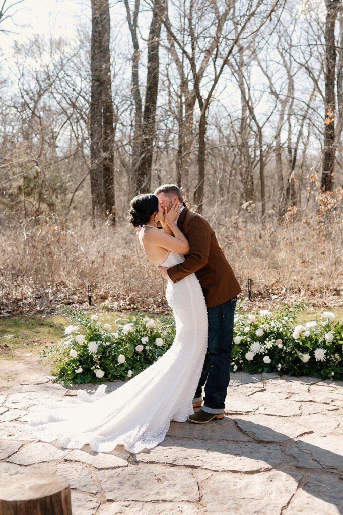 A bride and groom kiss outdoors during their microwedding near an earthy grounded floral arch.