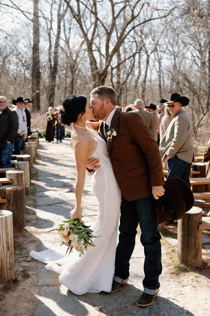 A groom kisses his bride in celebration of their outdoor microwedding at the end of a wedding aisle. 