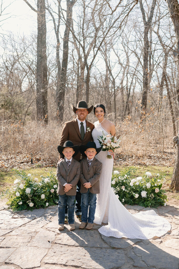 A bride and groom pose with two young family members in cowboy hats, suit jackets, cowboy boots and jeans. 