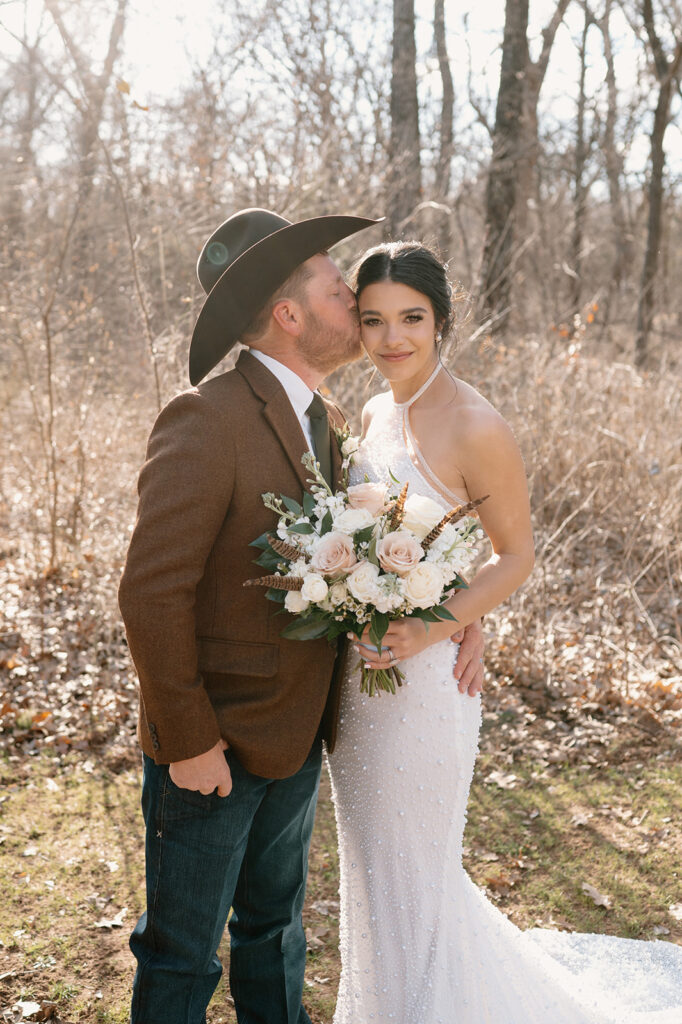 A man in a cowboy hat, brown suit jacket, tie and jeans kisses his bride on the forehead. 