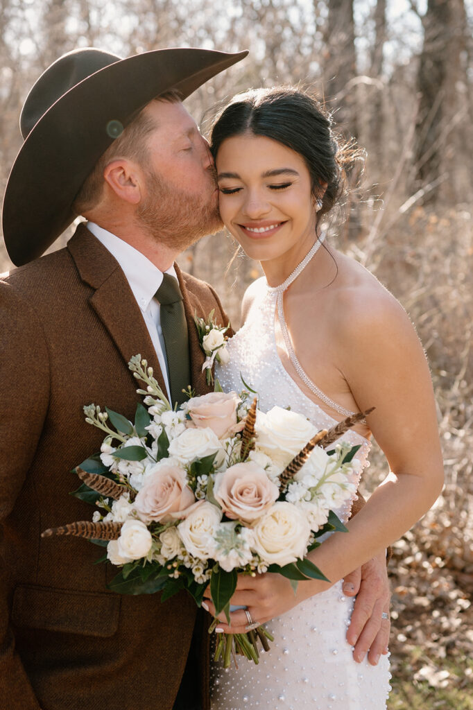 A groom kisses his bride's temple while she is holding a rose bridal bouquet filled with pink and white roses, feathers and greenary. 