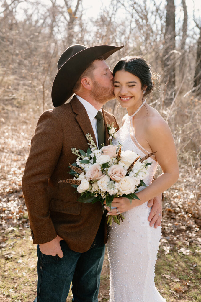 A groom kisses his bride as she smiles and holds a large rose bridal bouquet. 