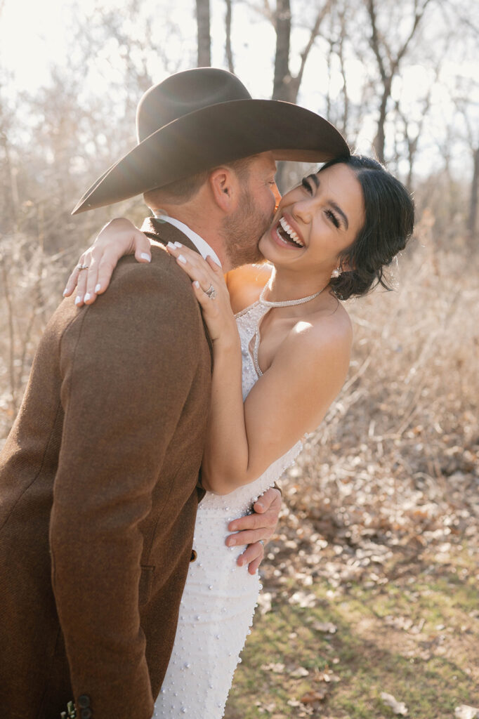 A man in a brown suit jacket and cowboy hat kisses his bride during their outdoor rustic microwedding.  