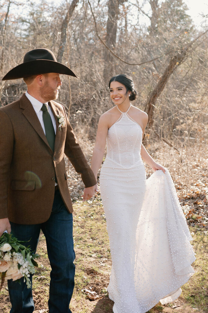 A bride and groom hold hands while walking through a forested area during their outdoor microwedding.