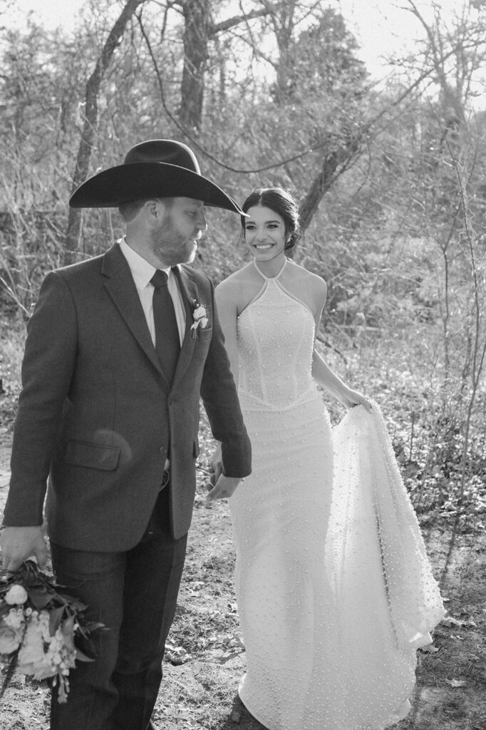 A groom holds a floral bouquet in one hand, while holding his bride's hand in another, walking through a forest. 