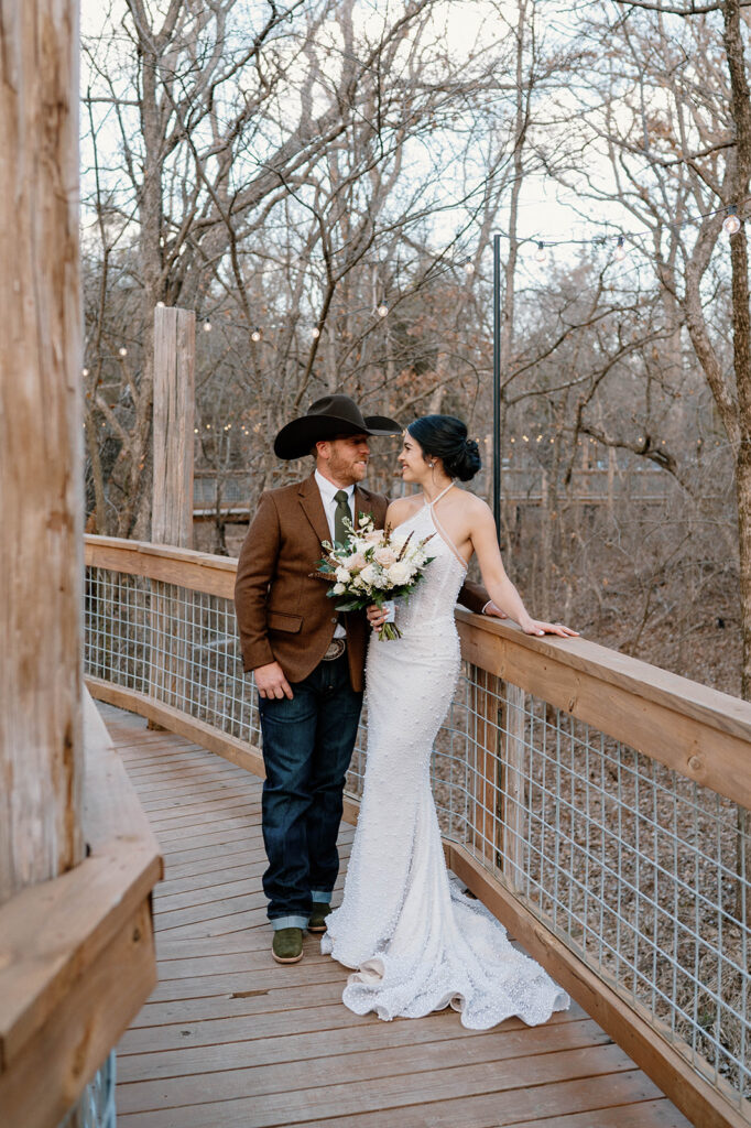 A bride leans on a wooden bride railing while smiling at her groom during their outdoor microwedding.