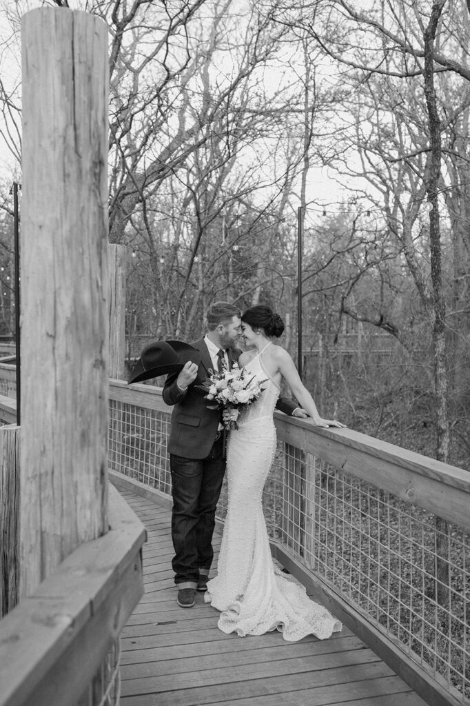 A man holds his cowboy hat while leaning in for a kiss towards his bride as they rest their hands on a railing of a wooden bridge. 