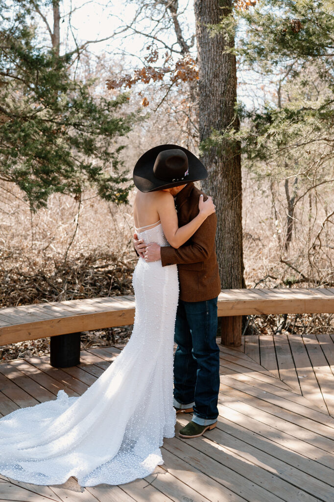 A bride in long, beaded white wedding dress hugs her groom who is wearing a cowboy hat, brown suit jacket, jeans and cowboy boots. 