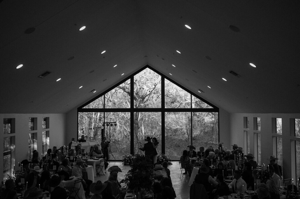 A bride and groom share a first dance in front of wedding guests in an intimate reception space with a large a-frame window. 