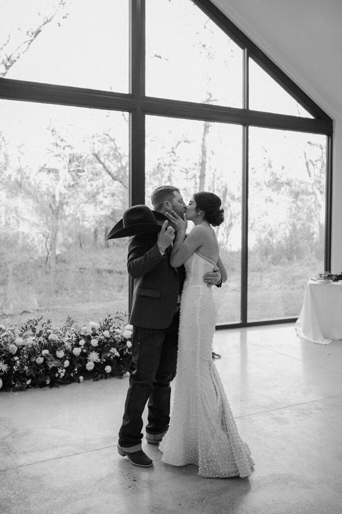 A bride and groom share a kisses near a gounded floral arch and large window at an indoor wedding reception. 