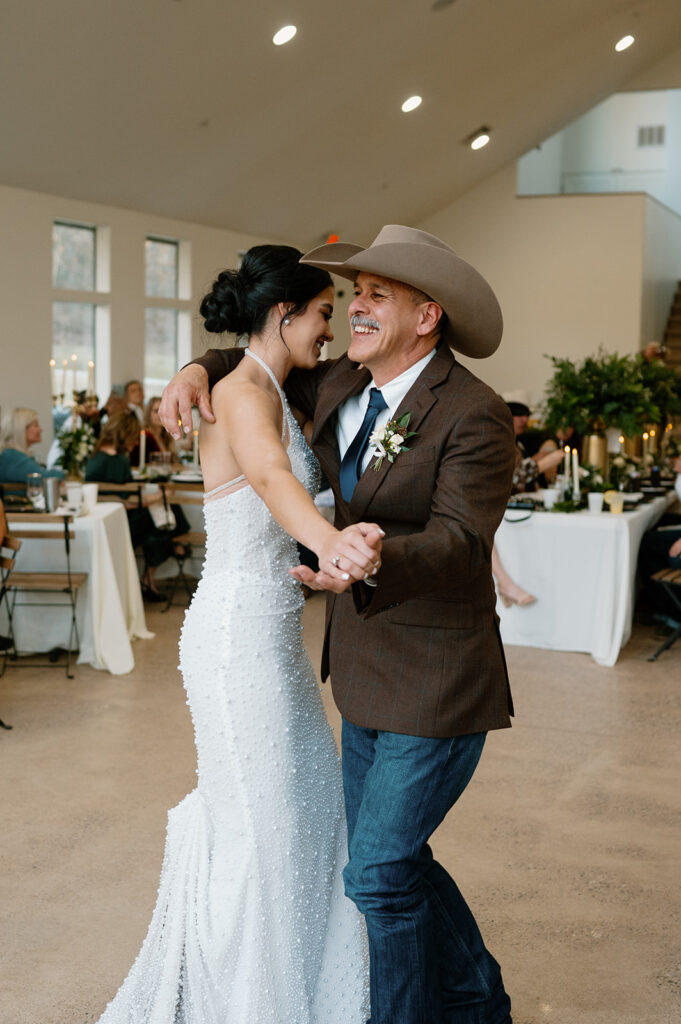 A father of the bride is wearing a cowboy hat, jeans and brown suit jacket while dancing with his daughter during a microwedding reception. 