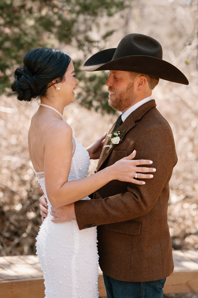 A bride with an updo is wearing a white beaded wedding gown and pearl earrings, while her groom is wearing a brown suit jacket, cowboy hat and dainty rose boutonniere.