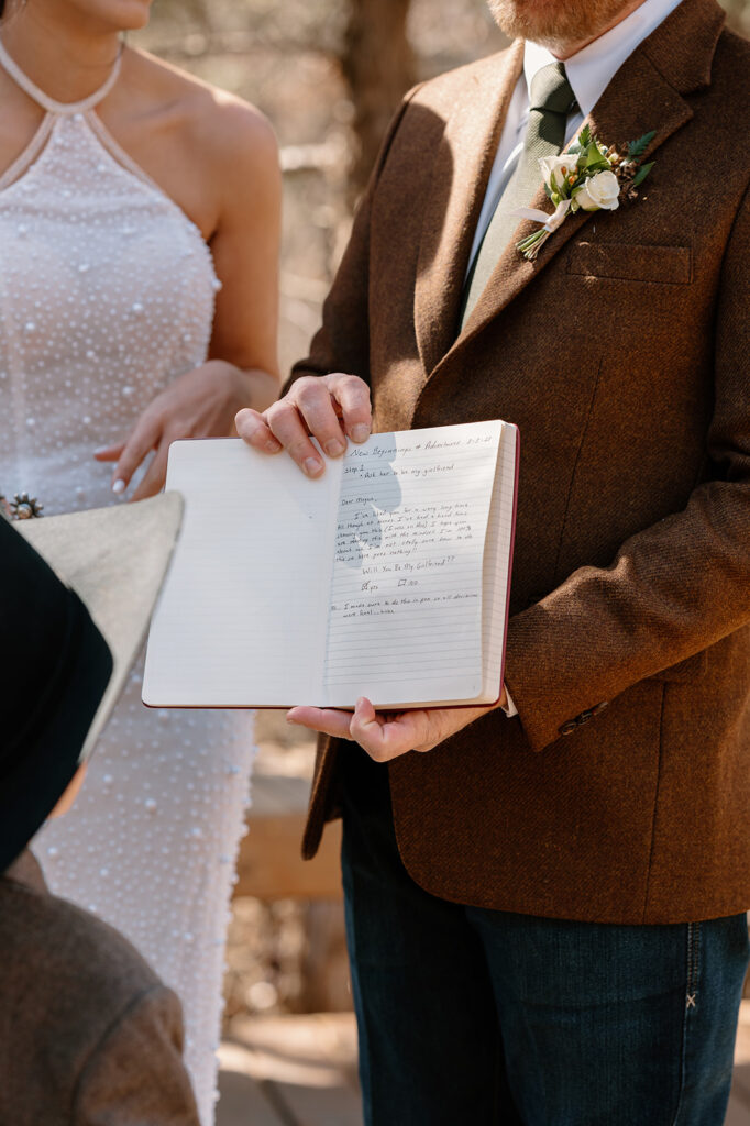 A groom in a brown suit jacket, decorated with a rose boutonniere on his lapel, holds a handwritten vow book during a microwedding ceremony.  