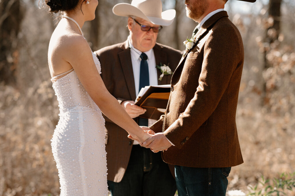 A bride, wearing a long, beaded wedding gown, holds her groom's hands, who is is wearing a brown blazer, cowboy hat and jeans. 