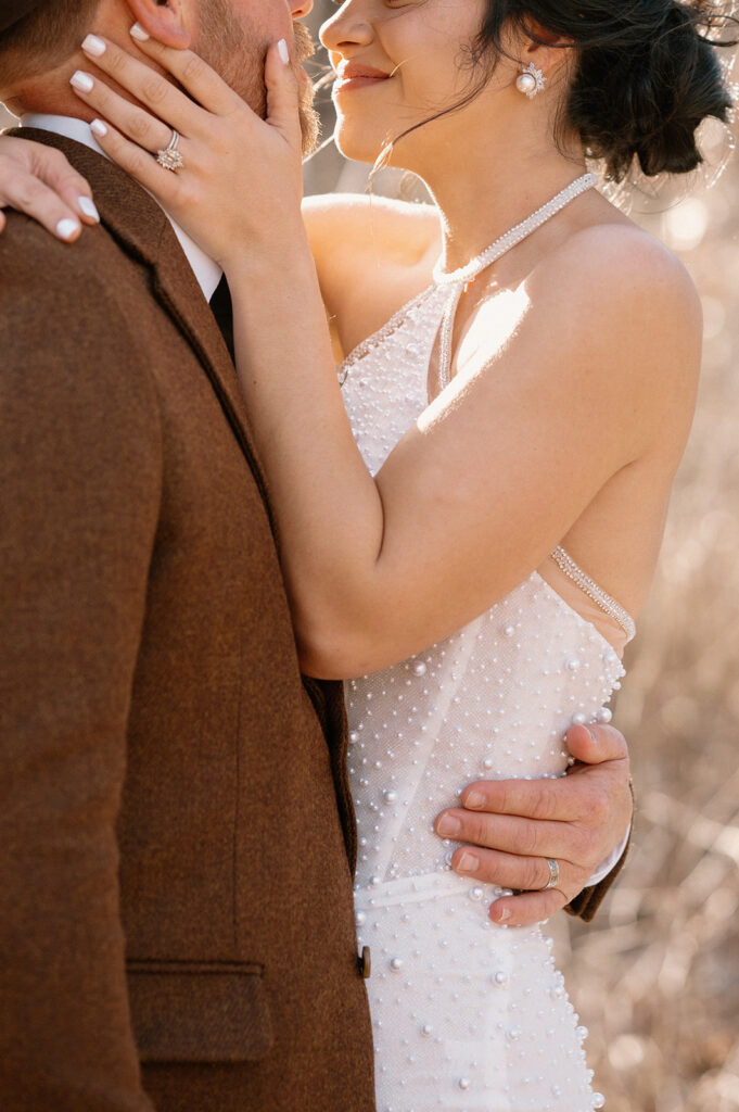 A bride wearing a large diamond ring holds her groom's face while taking outdoor wedding portraits. 