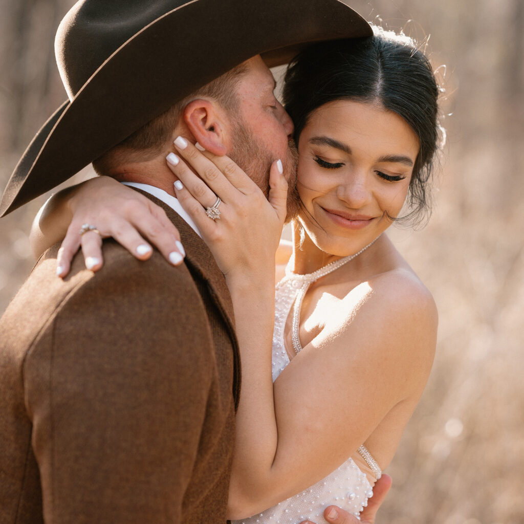 A man in a cowboy hat kisses his bride on the cheek during wedding portraits during their microwedding. 