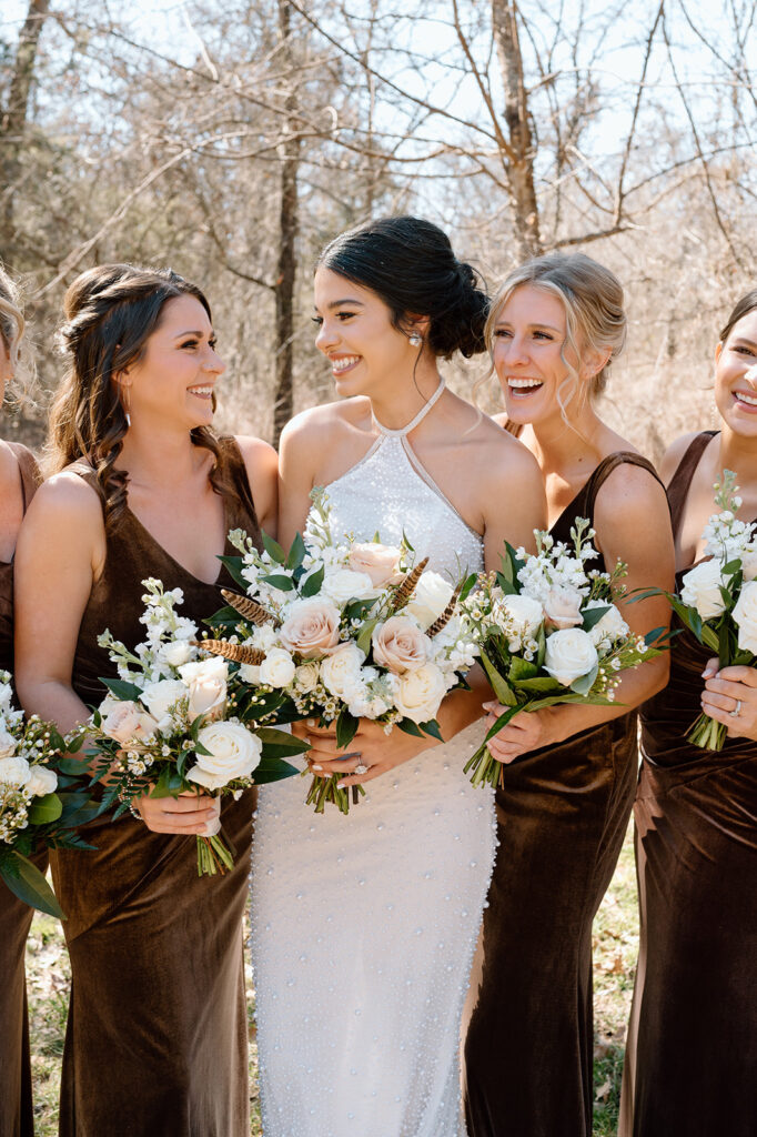 A bride, holding a rose bridal bouquet with feathers, poses with bridesmaids, who are all wearing brown velvet bridesmaid dress.