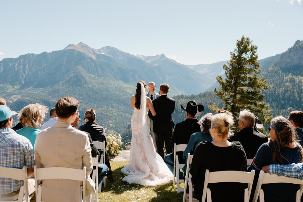 Wedding guests watch as a  bride and groom recite vows with an officiant during an outdoor ceremony overlooking Ouray at Gold Mountain Ranch.