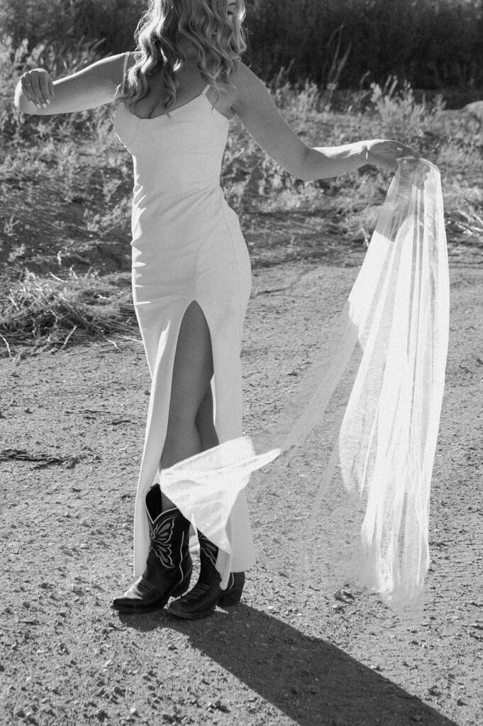 A bride in a floor length wedding gown and cowboy boots dances along a dirt road while holding her veil. 