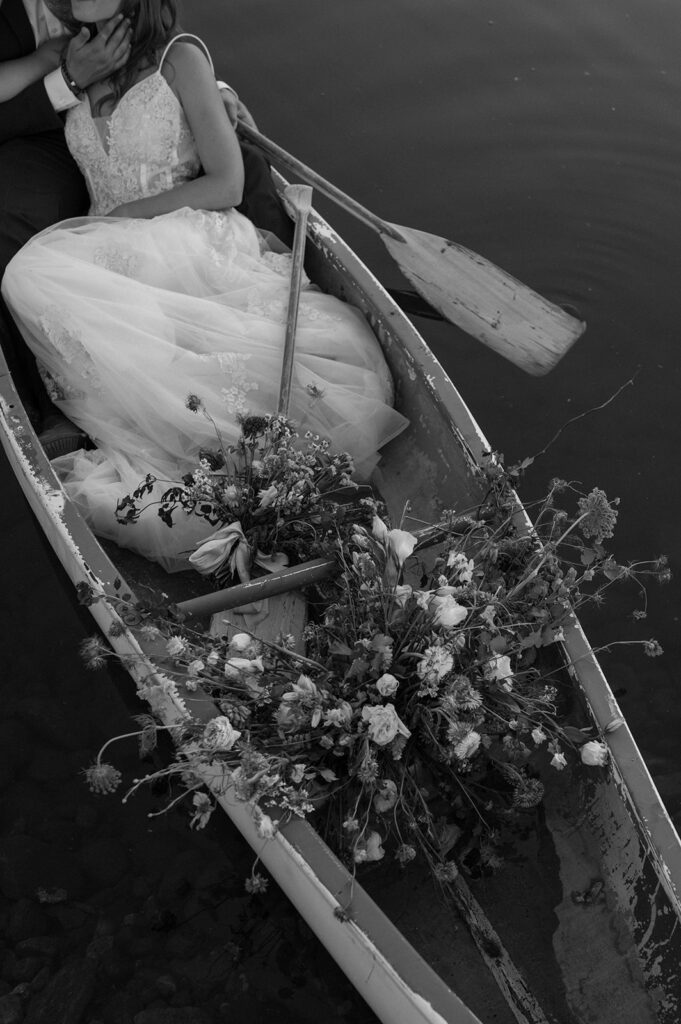 A bride in a long, lace wedding gown is seen lying in a row boat on a pond with her groom and large bridal bouquet at Shaw's River Ranch.