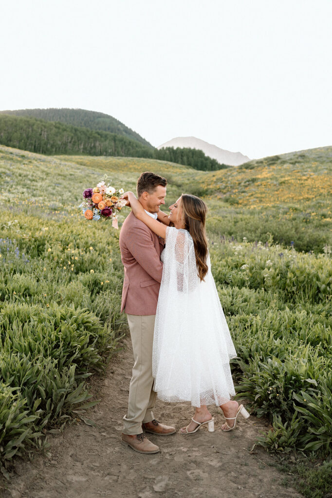 A bride in a ankle-length wedding dress and block heels embraces her groom, who is wearing a dusty rose suit coat, along a Crested Butte wildflower hike.