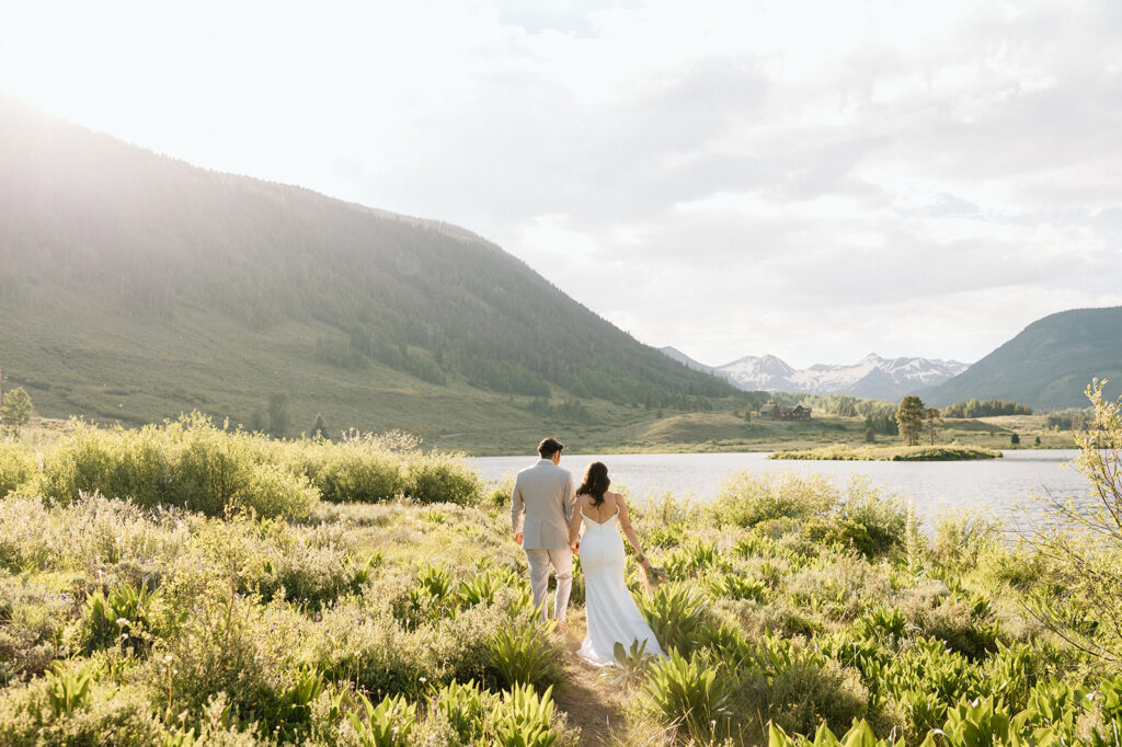 A bride and groom walk to a water's edge with the Rocky Mountains in the distance during their Colorado mountain elopement. 