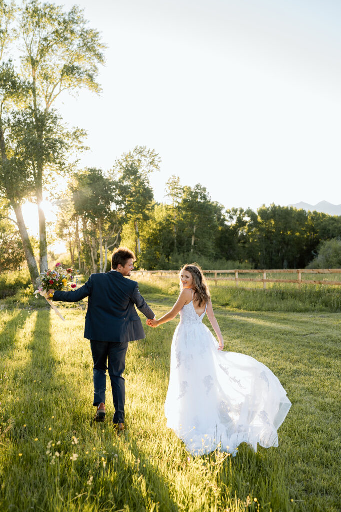 A bride in a long white wedding gown holds her groom's hand while walking through a grassy field on Shaw's River Ranch.