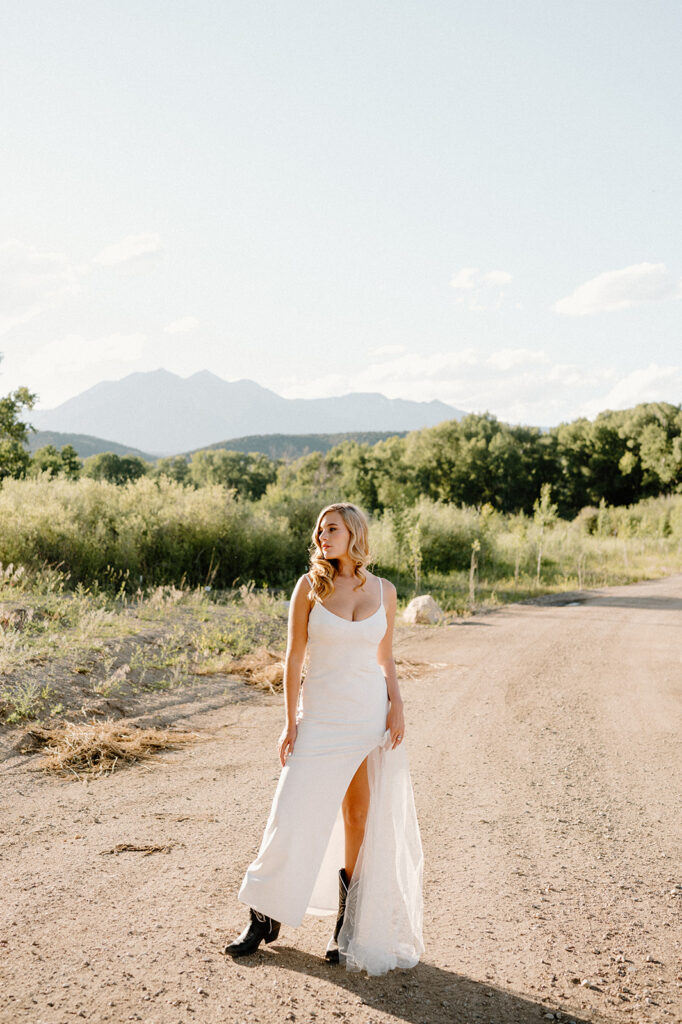 A bride in a long, scoop neck wedding dress with spaghetti straps poses along a dirt road in the Rockies during her Colorado intimate wedding. 