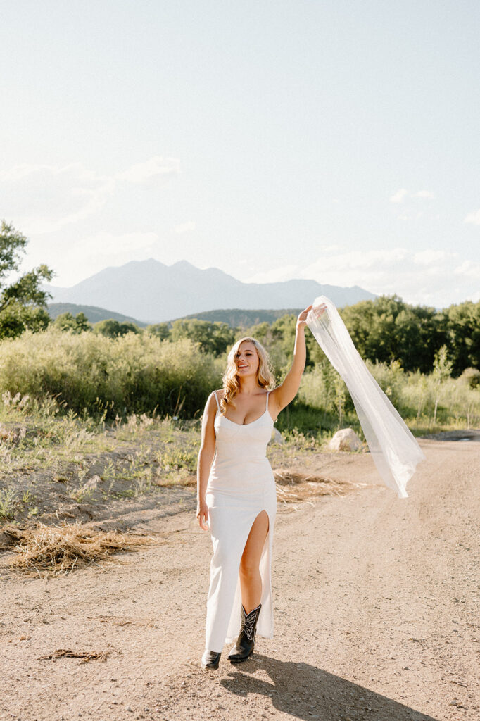 A bride in a long, scoop neck wedding dress with spaghetti straps walks along a dirt road in the Rockies during her Colorado intimate wedding. 