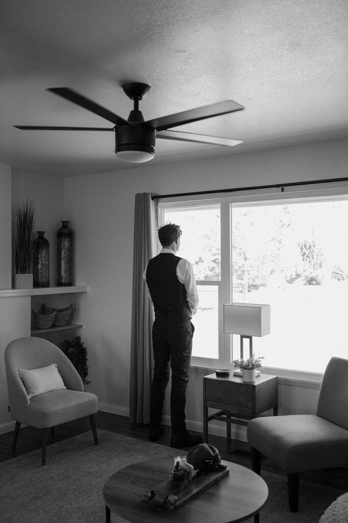 A groom in a wedding tux looks out the window of a Moab Airbnb. 