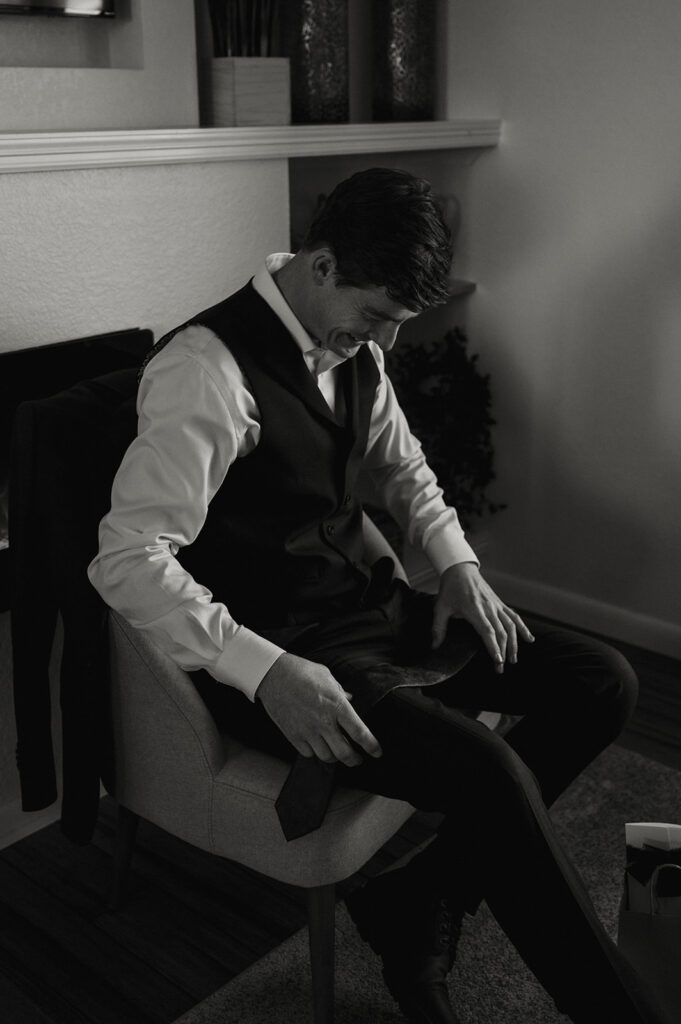 A groom admires his tie while sitting in a chair at a Moab Airbnb.