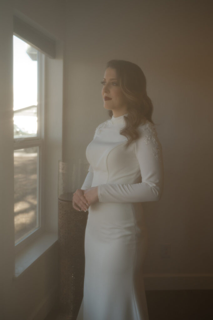 A bride in a long sleeve wedding gown looks out the window of a Moab Airbnb. 