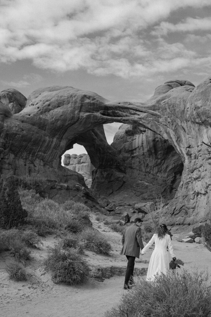 A Moab elopement couple holds hands while walking towards Double Arches in Arches National Park.