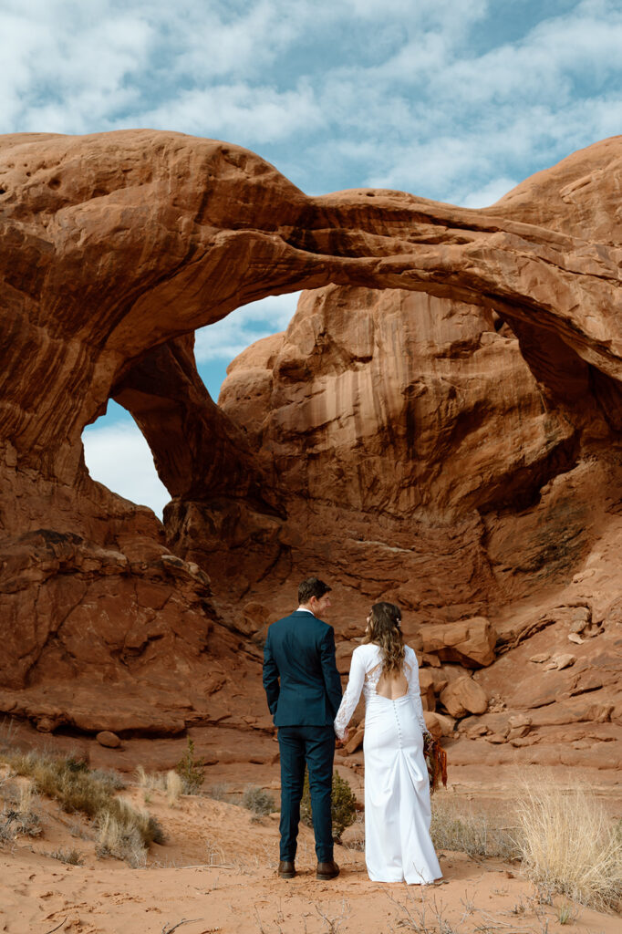 A Utah couple holds hands while looking at one another in front of Double Arches in Arches National Park.