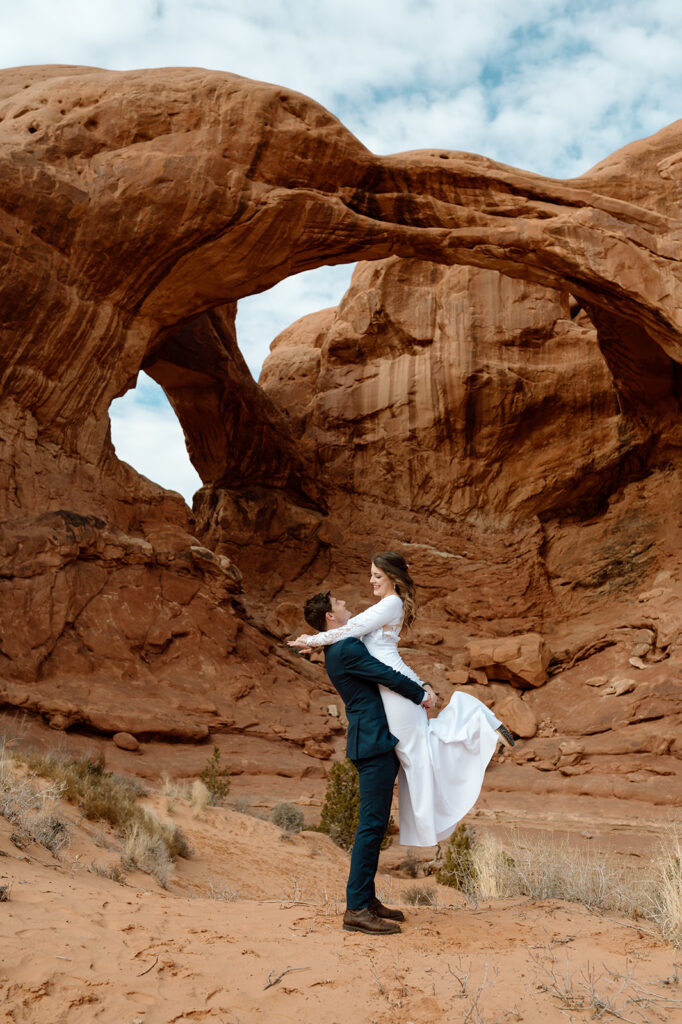 A groom in a navy wedding suit and brown boots lifts up bride near Double Arches in Arches National Park.