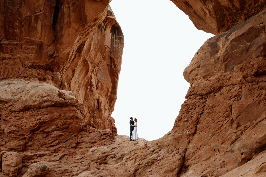 A couple stands beneath a red rock arch for wedding portraits during their Arches National Park elopement. 