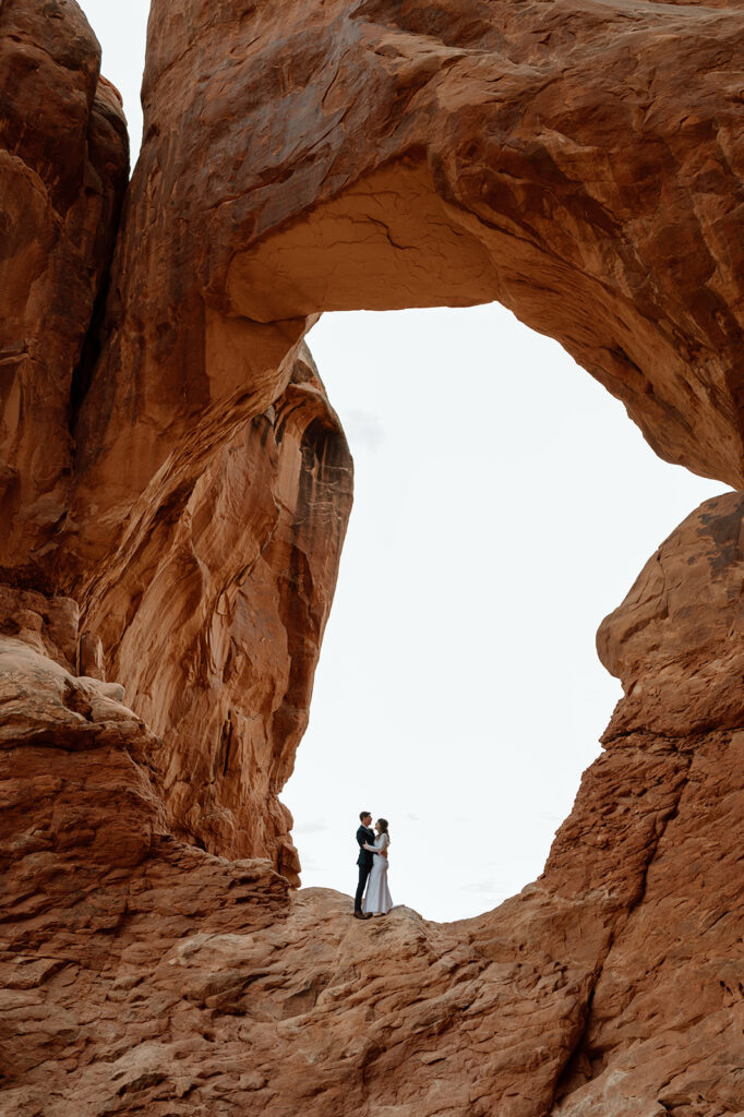 A couple stands in a red rock arch in Arches National Park during their Moab elopement in the desert. 