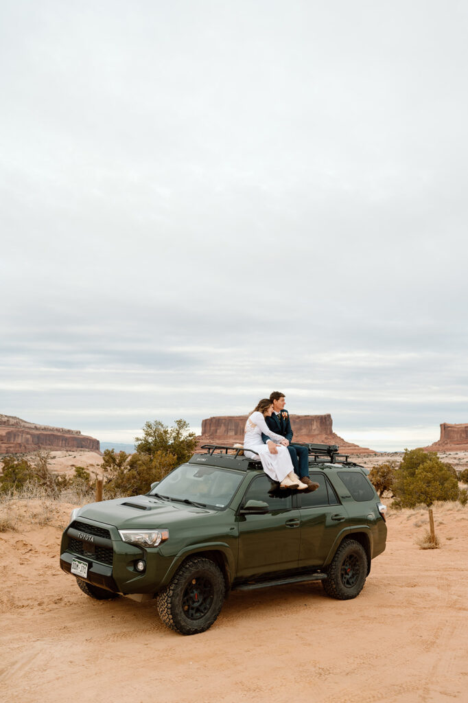 A Moab elopement couple sits atop a dark green 4x4 vehicle during their desert elopement. 