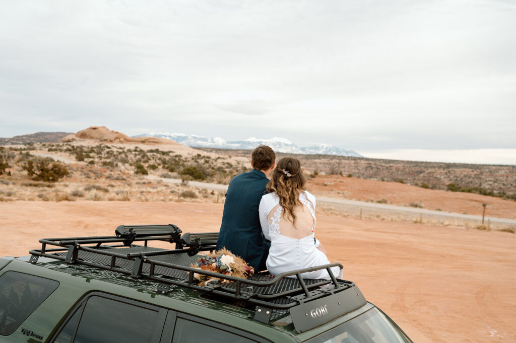 A Utah elopement couple admires snowy mountain peaks in the distance while sitting on the roof of a 4x4 vehicle. 