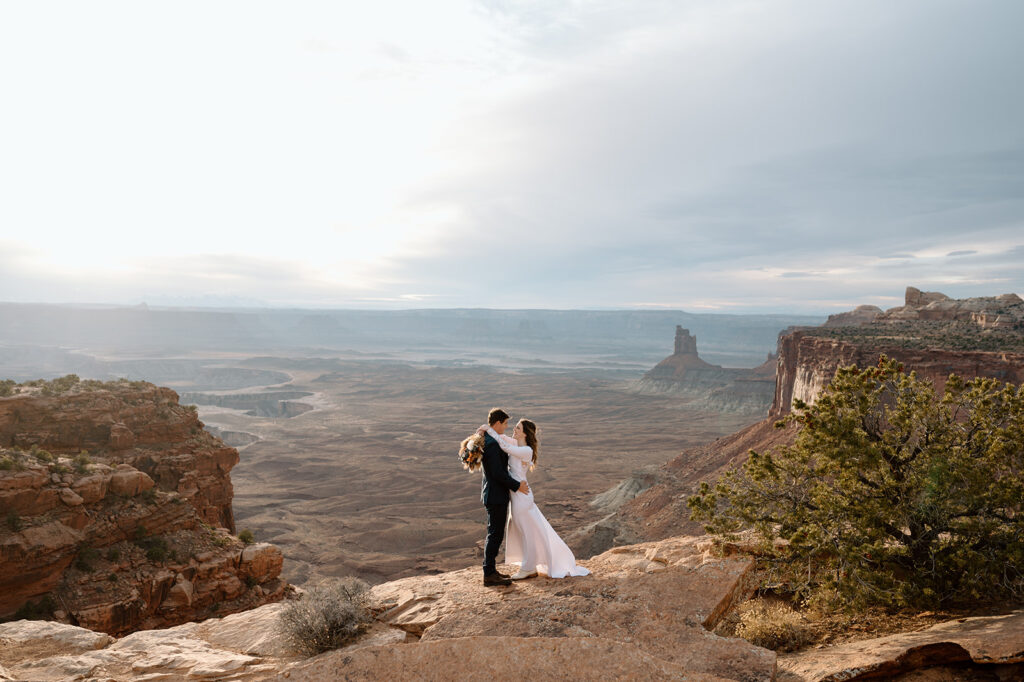 A man, wearing a navy suit and a woman, wearing a long sleeve wedding dress and holding a dried floral bouquet, embraces on an overlook in Canyonlands National Park.