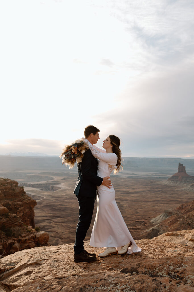 A groom in a navy tux embraces his wife, who is dressed in a long white wedding dress and white boots, on a cliff in Canyonlands. 