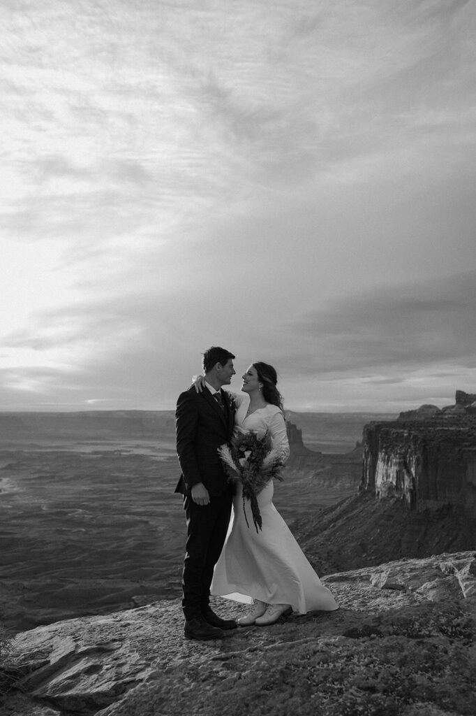 A bride admires her groom while taking Utah wedding portraits on a cliff in Canyonlands National Park. 